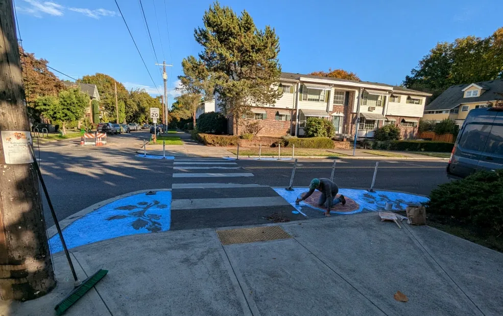 A neighbor kneels to paint a terracotta circle on a bright blue curb extension at NE 21st and Clackamas. Dark leaf shapes cover the blue, white flexible posts edge both painted corners, and a zebra crosswalk runs between them. A push broom leans against a utility pole in the foreground.